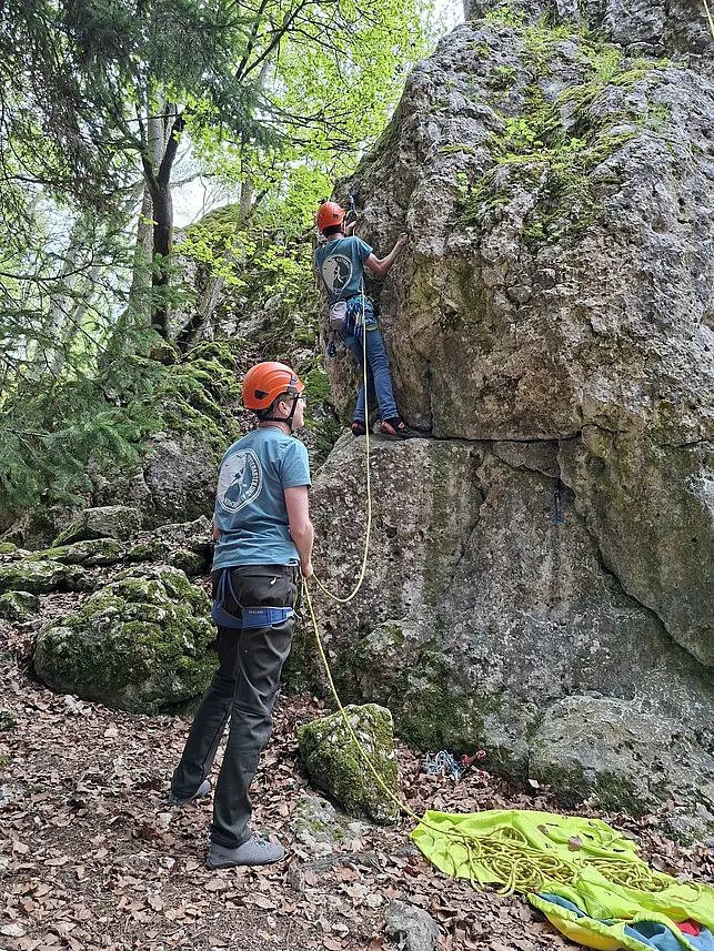 Zwei Personen mit Kletterausrüstung klettern an einem Felsen im Wald. Eine Person sichert die andere mit einem Seil, während die zweite Person am Felsen hochklettert. Beide tragen Helme und ähnliche Kleidung.