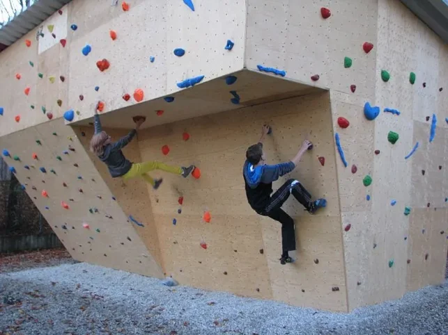 Zwei Personen klettern an einer künstlichen Kletterwand mit bunten Griffen im Freien. Die Wand ist überhängend, und beide nutzen Hände und Füße, um sich daran festzuhalten.