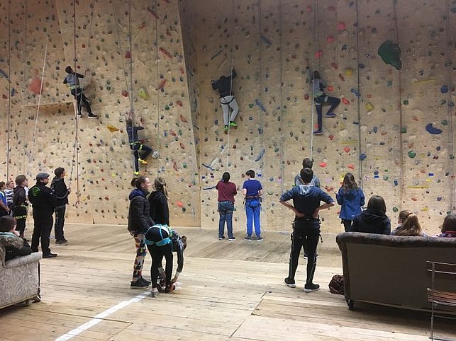 Mehrere Personen klettern an einer hohen Indoor-Kletterwand mit vielen bunten Griffen, während andere am Boden zuschauen oder sich vorbereiten. Die Atmosphäre wirkt sportlich und gemeinschaftlich.