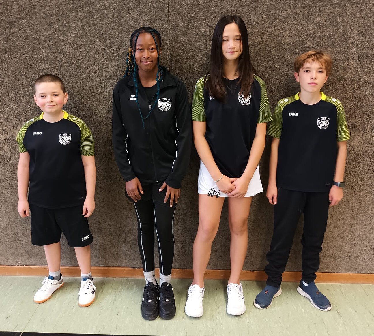 Vier Kinder in Sportkleidung stehen nebeneinander in einer Turnhalle vor einer Wand und schauen in die Kamera. Sie tragen dunkle Trikots mit einem Vereinslogo.