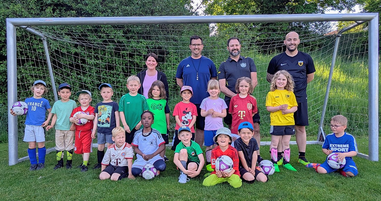 Eine Kinderfußballmannschaft mit mehreren Kindern und vier Erwachsenen posiert gemeinsam vor einem Fußballtor auf einem grünen Sportplatz. Einige Kinder halten Fußbälle in den Händen und alle lächeln in die Kamera.