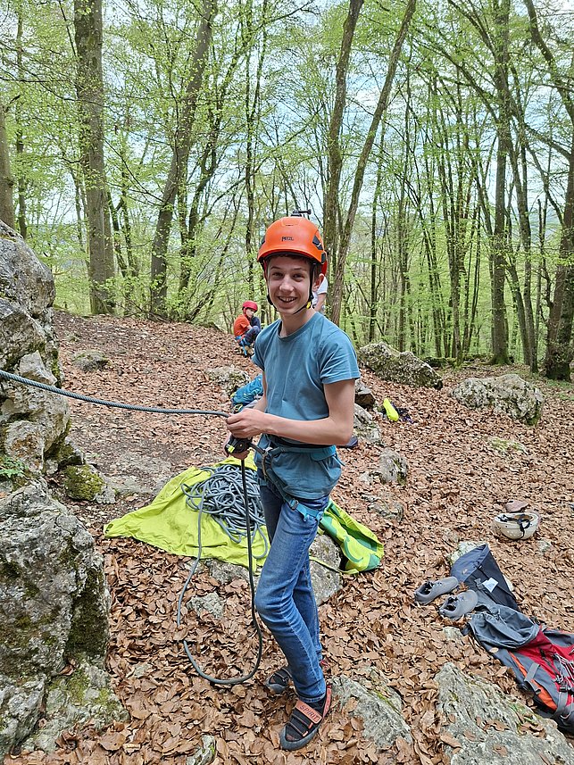 Ein Jugendlicher mit Kletterhelm steht im Wald und hält ein Kletterseil. Er lächelt und bereitet sich auf das Klettern vor. Im Hintergrund sind Bäume und Kletterausrüstung.