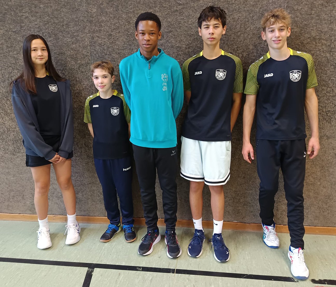Fünf Jugendliche stehen nebeneinander in einer Sporthalle, tragen Sportkleidung und schauen in die Kamera. Drei Jungen und ein Mädchen tragen dunkle Trikots mit Vereinslogo, einer trägt einen türkisfarbenen Pullover.