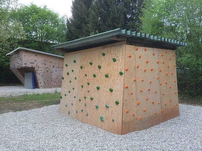 Zwei Holzwände im Freien sind mit bunten Klettergriffen ausgestattet und dienen als Boulderwände zum Klettern. Die Umgebung ist grün und bewaldet.