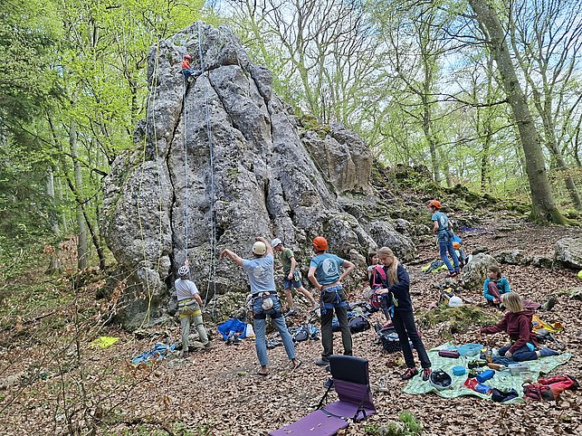 Mehrere Menschen klettern und sichern an einem großen Felsen im Wald. Einige Personen sitzen oder stehen am Boden und beobachten die Kletternden. Kletterausrüstung ist sichtbar.
