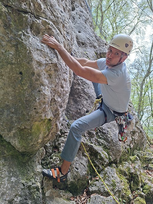 Ein Mann mit Kletterausrüstung und Helm klettert an einer steilen Felswand im Freien. Er hält sich mit Händen und Füßen fest und ist durch ein Seil gesichert.