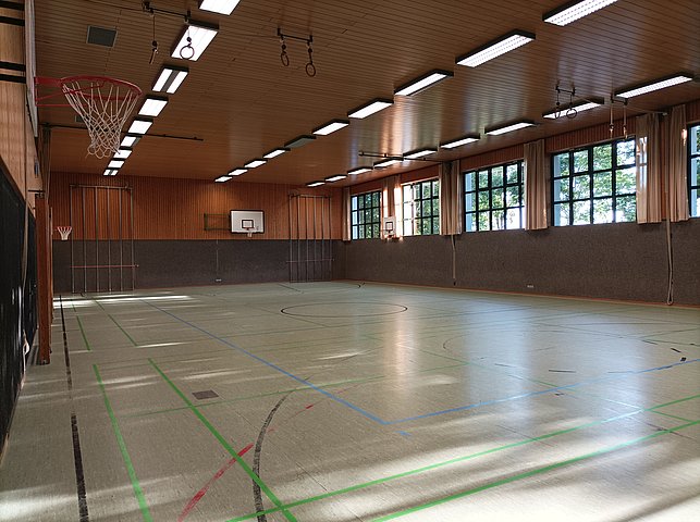 Eine leere Turnhalle mit Basketballkörben an beiden Enden, vielen Fenstern an der Seite und Spielfeldmarkierungen auf dem Boden.