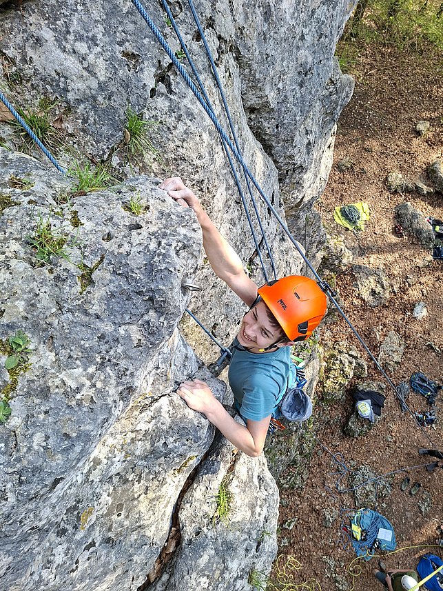 Ein junger Mann mit orangefarbenem Helm klettert an einer steilen Felswand nach oben und ist mit Seilen gesichert. Die Szene vermittelt Klettersport und Konzentration.
