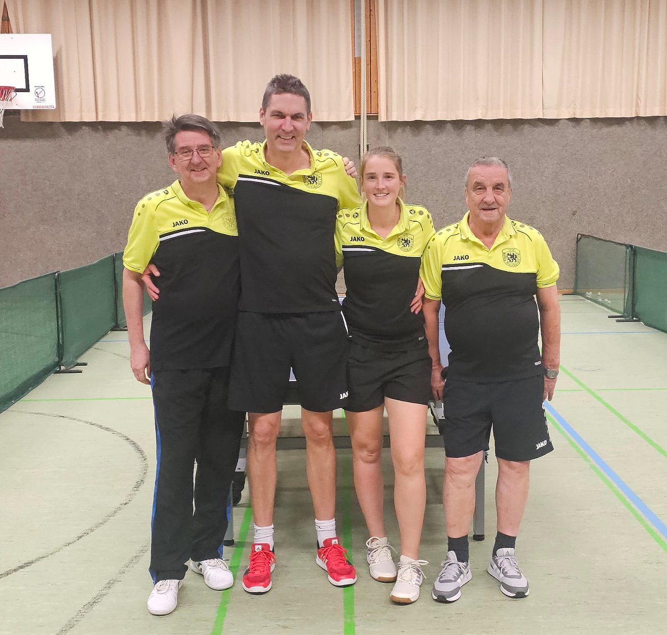 Vier Tischtennisspieler stehen in einer Sporthalle nebeneinander und lächeln in die Kamera. Sie tragen einheitliche gelbe und schwarze Sportkleidung mit Vereinslogo. Im Hintergrund ist ein Tischtennistisch und eine Turnhallenwand zu sehen.