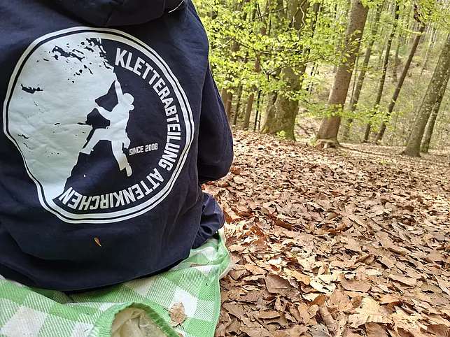 Eine Person sitzt auf einer Decke im Wald. Auf ihrer Jacke ist ein großes Logo mit einer Kletterfigur und dem Schriftzug „Kletterabteilung Altenkirchen“. Um sie herum liegen viele braune Blätter auf dem Waldboden.