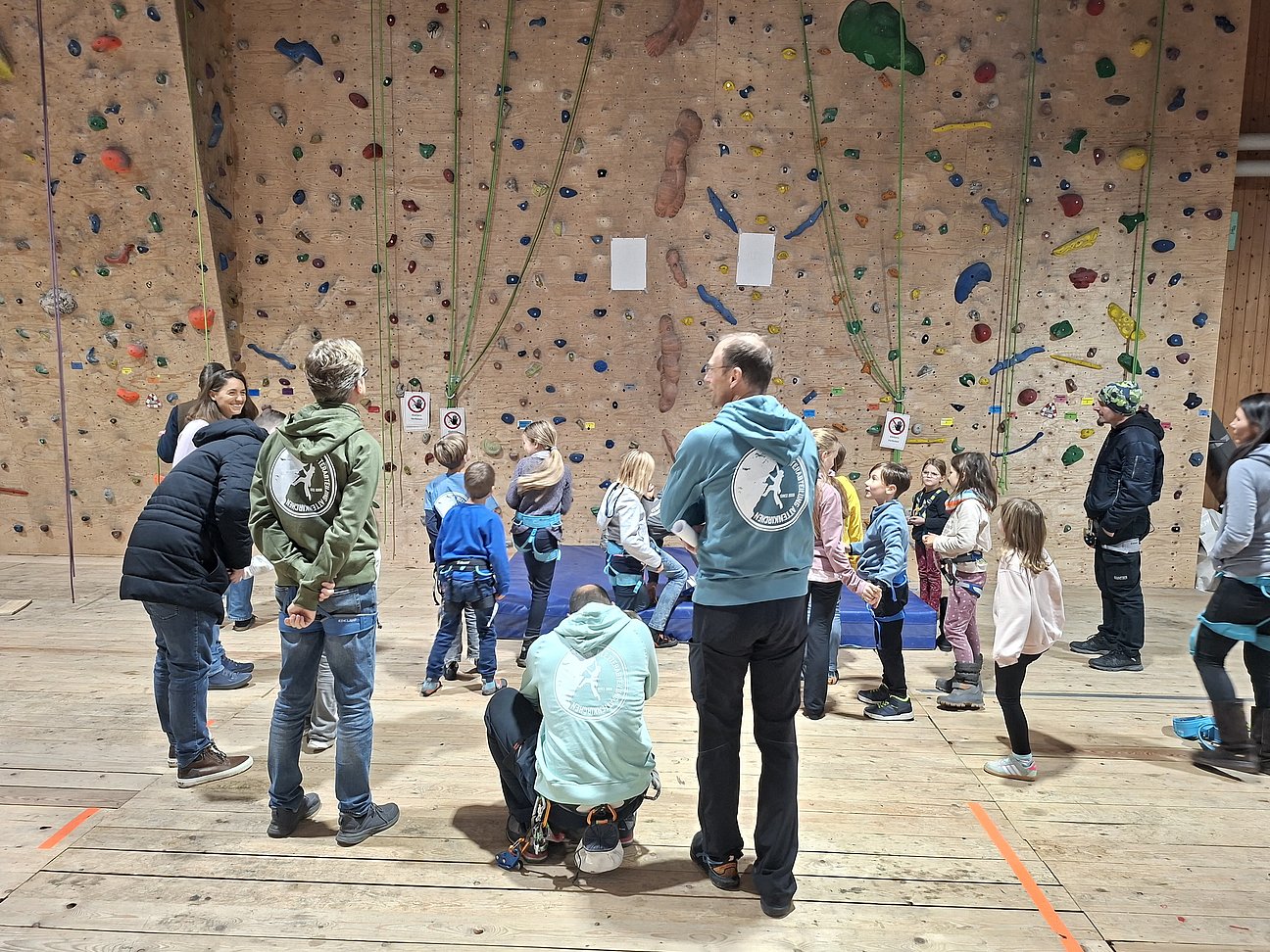 Mehrere Kinder und Erwachsene versammeln sich in einer Kletterhalle vor einer großen Kletterwand, als Vorbereitung auf das Klettern. Die Kinder tragen Klettergurte und werden offenbar von Erwachsenen betreut oder unterrichtet.