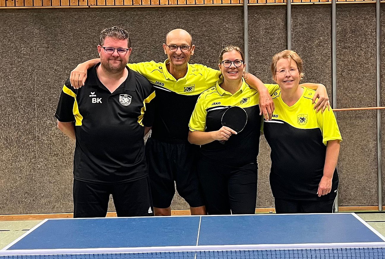 Vier Tischtennisspielerinnen und -spieler stehen gemeinsam hinter einer blauen Tischtennisplatte in einer Sporthalle und lächeln in die Kamera. Sie tragen schwarz-gelbe Sportkleidung.