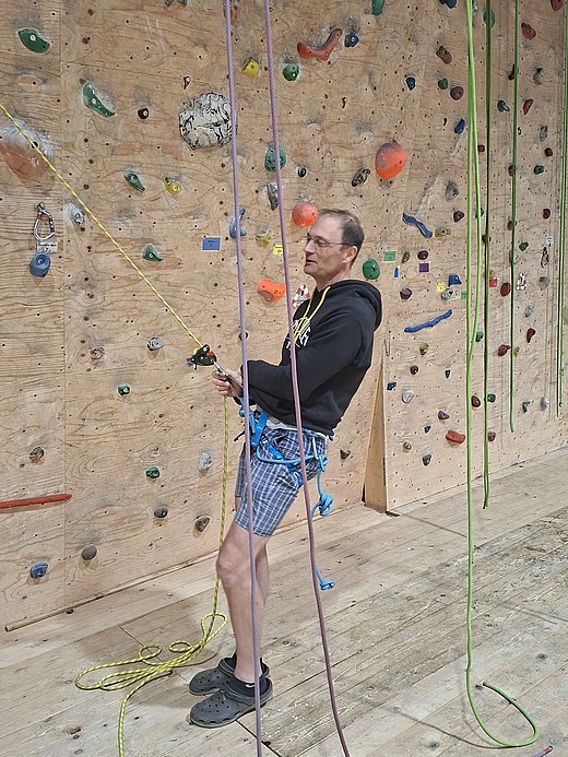 Ein Mann sichert eine Kletterin oder einen Kletterer in einer Kletterhalle. Er trägt einen Klettergurt und hält ein Seil an einer Kletterwand mit bunten Griffen.