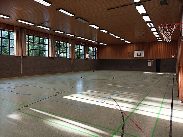 Eine leere Sporthalle mit mehreren Basketballkörben, großen Fenstern entlang einer Wand und markierten Spielfeldlinien auf dem Boden.