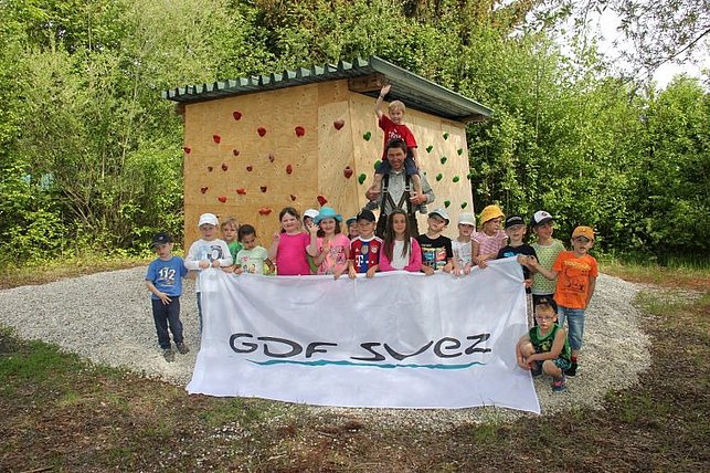 Eine Gruppe von Kindern und einem Erwachsenen steht fröhlich vor einer kleinen Holzhütte mit Klettergriffen im Grünen und hält ein weißes Banner mit der Aufschrift "GDF SUEZ".