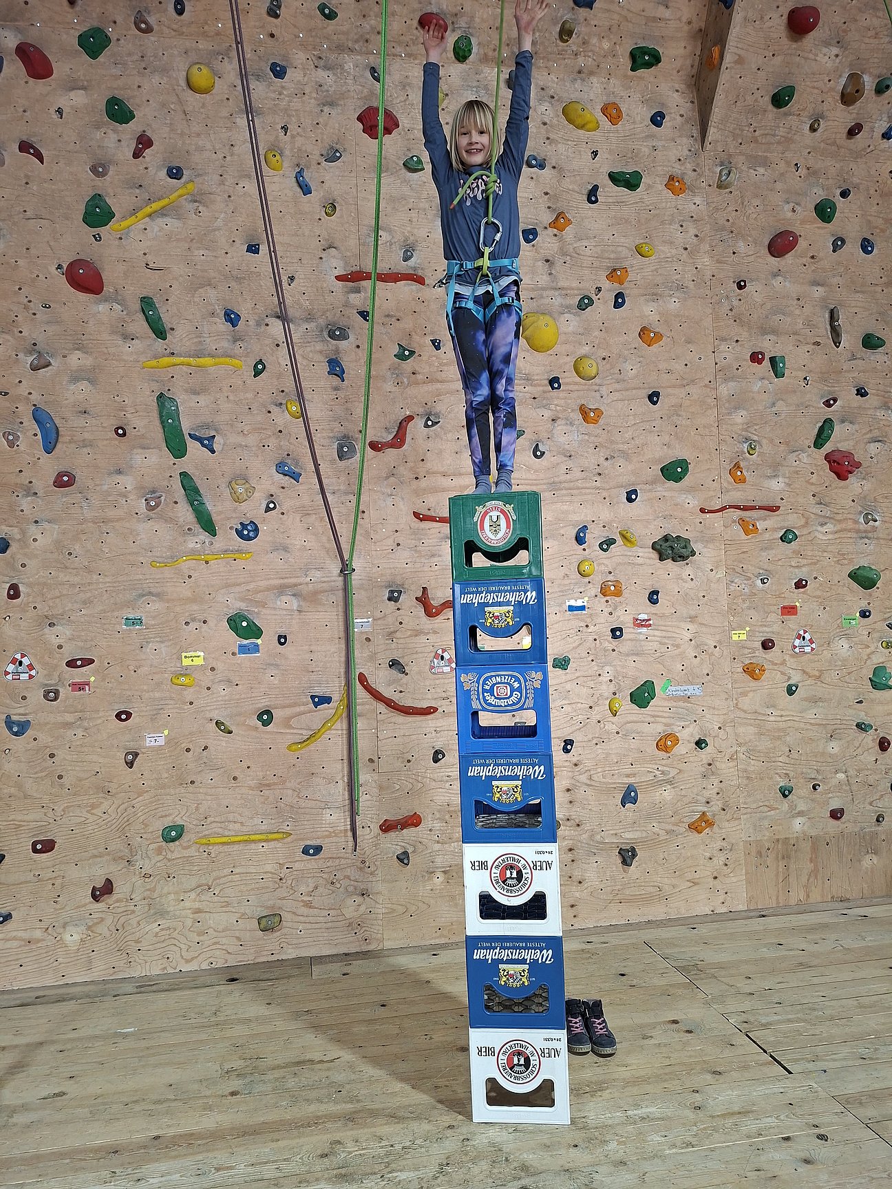 Ein Kind steht gesichert auf einem hohen Turm aus Getränkekisten in einer Kletterhalle. Im Hintergrund ist eine Kletterwand mit bunten Griffen zu sehen.
