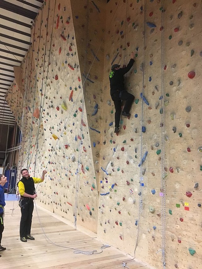 Zwei Personen beim Klettern in einer Halle: Eine Person klettert an einer Indoor-Kletterwand, während die andere Person sie am Boden sichert. Die Wand ist mit vielen bunten Griffen und Tritten ausgestattet.