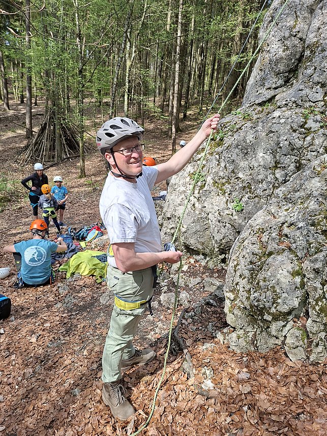 Ein Mann mit Helm und Klettergurt sichert eine Person beim Klettern an einem Felsen im Wald. Im Hintergrund sind mehrere weitere Kletterer.