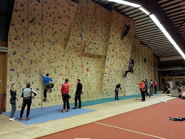 Mehrere Personen klettern an einer großen künstlichen Kletterwand in einer Sporthalle, während andere am Boden sichern oder zusehen. Die Szene zeigt einen belebten Indoor-Kletterbereich mit unterschiedlichen Schwierigkeitsgraden und aktiver Teilnahme.