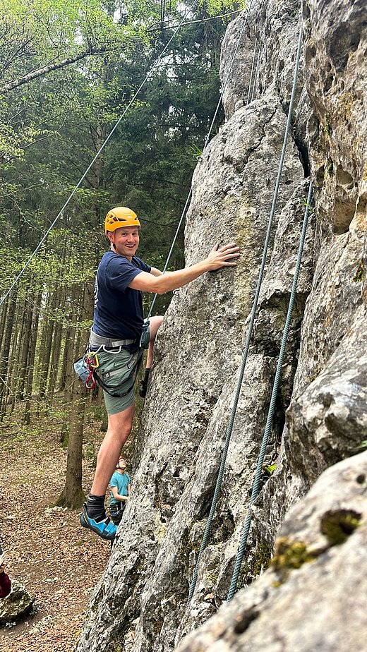Ein Mann mit Kletterausrüstung klettert an einer steilen Felswand im Wald. Er trägt einen Helm und hält sich mit einer Hand am Felsen fest. Kletterseile verlaufen neben ihm die Wand hinauf.