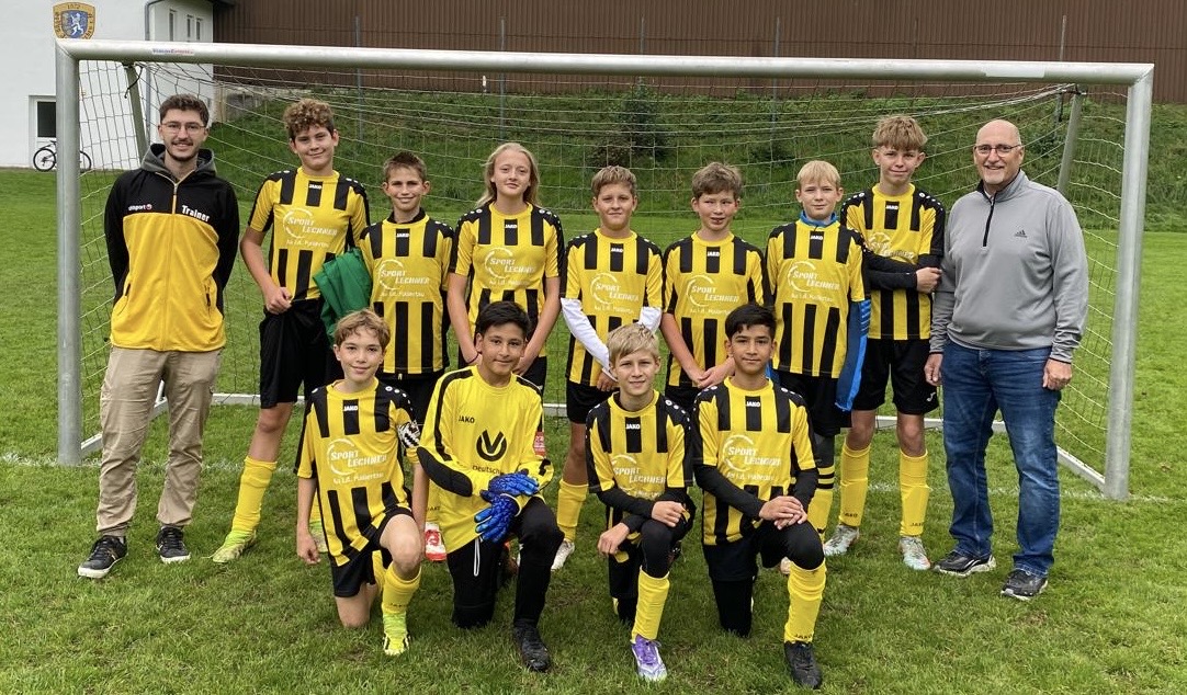Eine Fußballmannschaft mit Jugendlichen in gelb-schwarzen Trikots und zwei Trainern steht vor einem Tor auf einem Fußballfeld und posiert für ein Mannschaftsfoto.