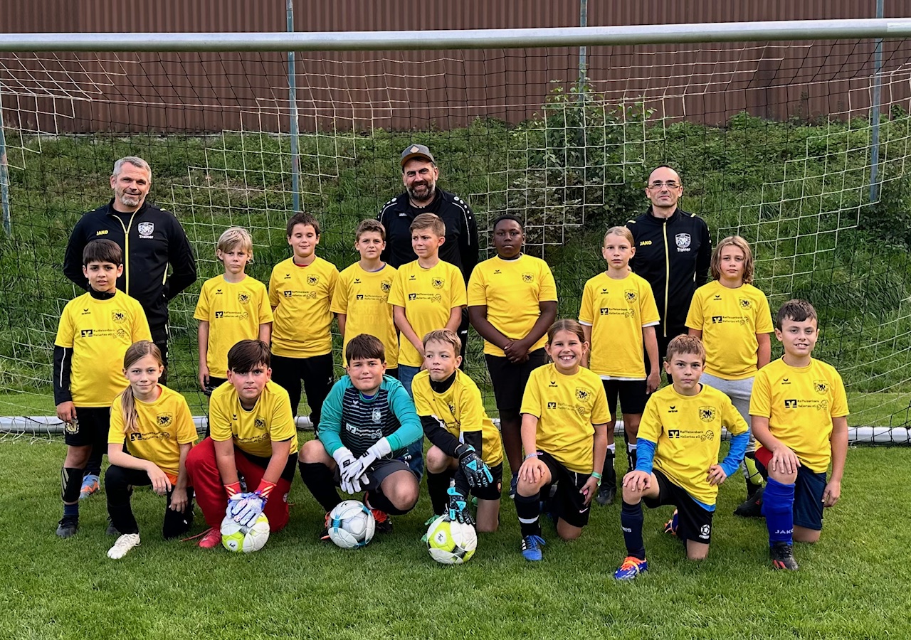 Eine Jugend-Fußballmannschaft mit drei Trainern steht und kniet gemeinsam vor einem Fußballtor. Die Kinder tragen gelbe Trikots, einige halten Fußbälle. Alle blicken in die Kamera und posieren für ein Mannschaftsfoto auf dem Fußballfeld.